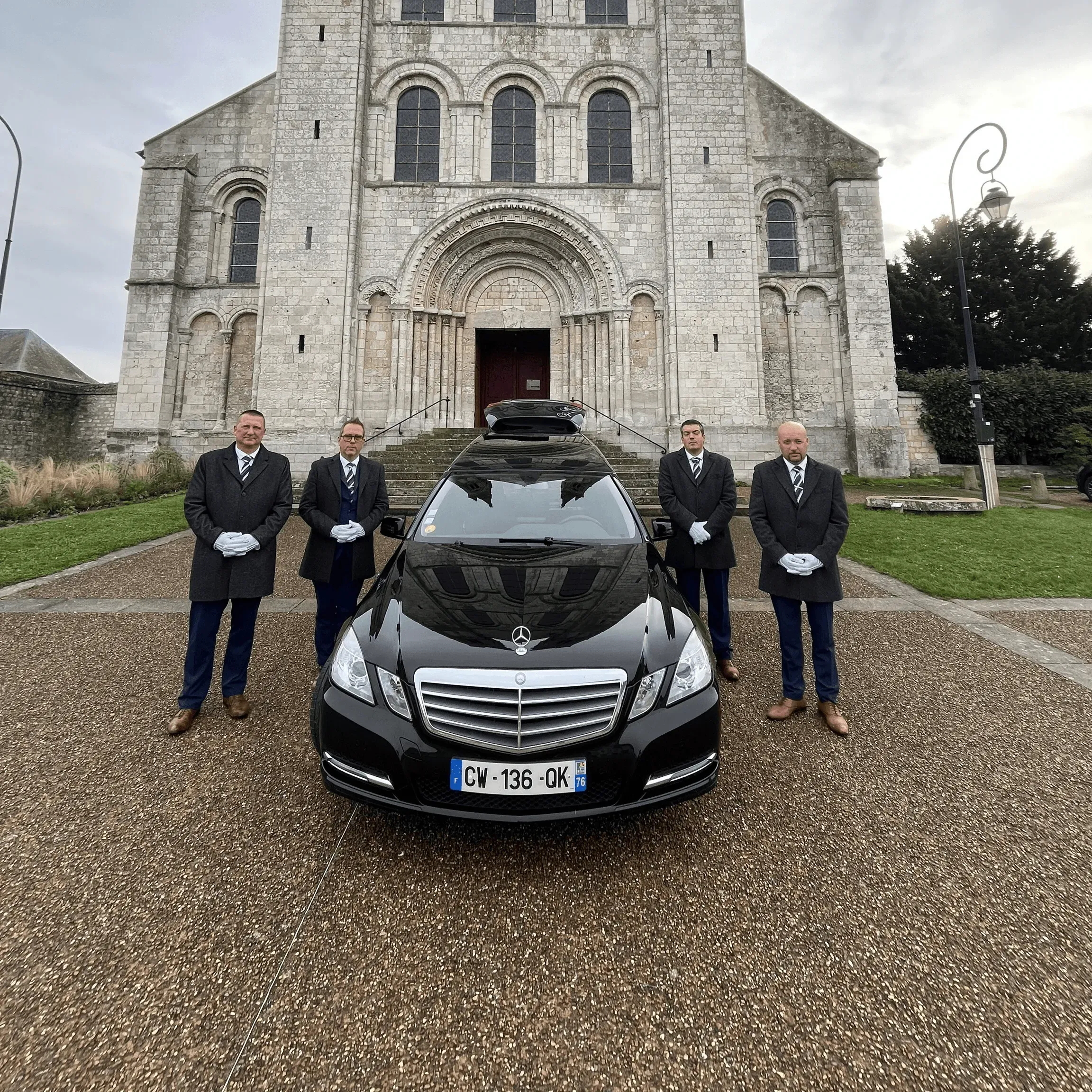 Porteurs funéraires en livrée devant un corbillard Mercedes et une église en pierre.