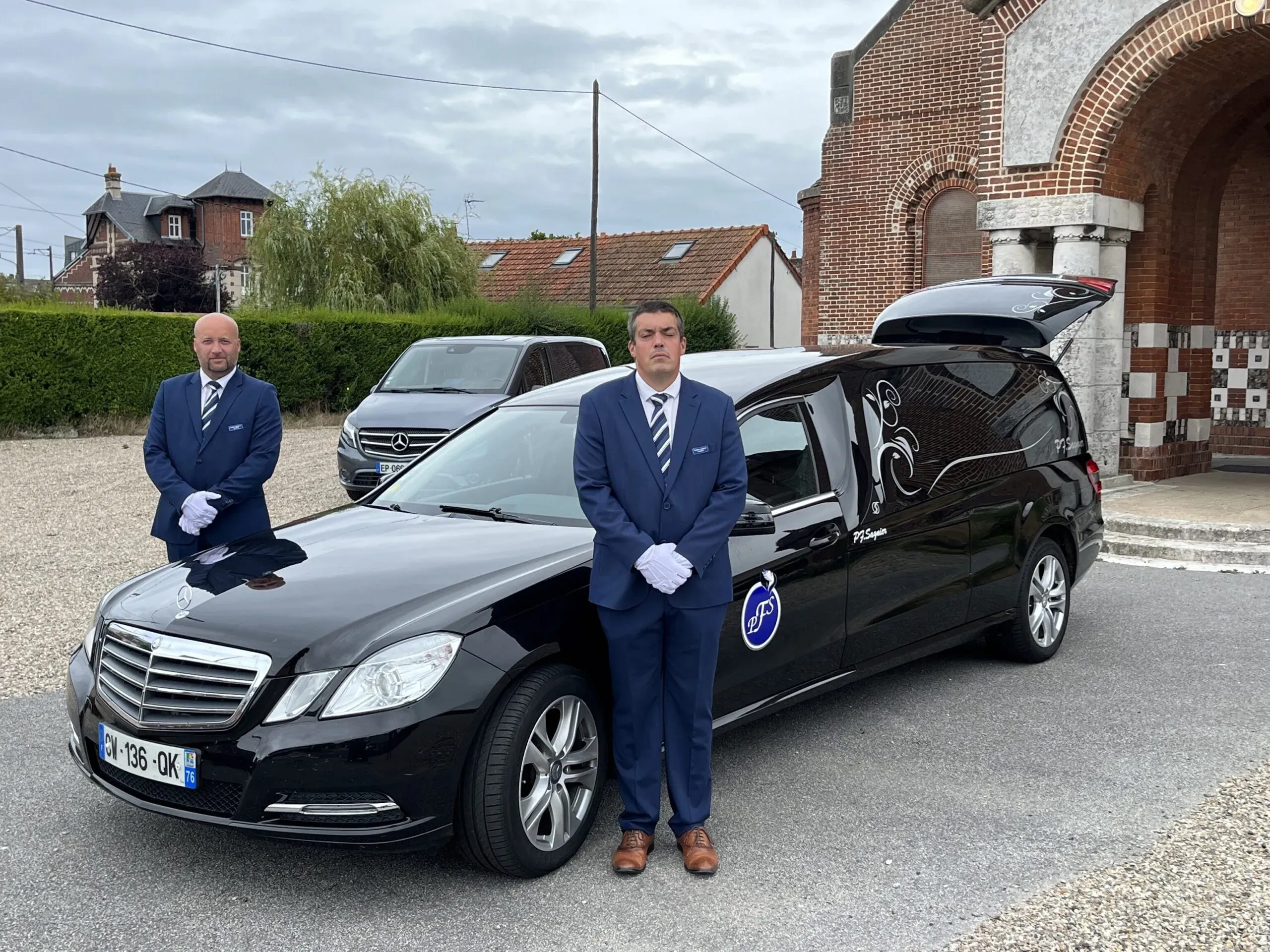 Deux porteurs funéraires en uniforme bleu posant devant un corbillard Mercedes noir.