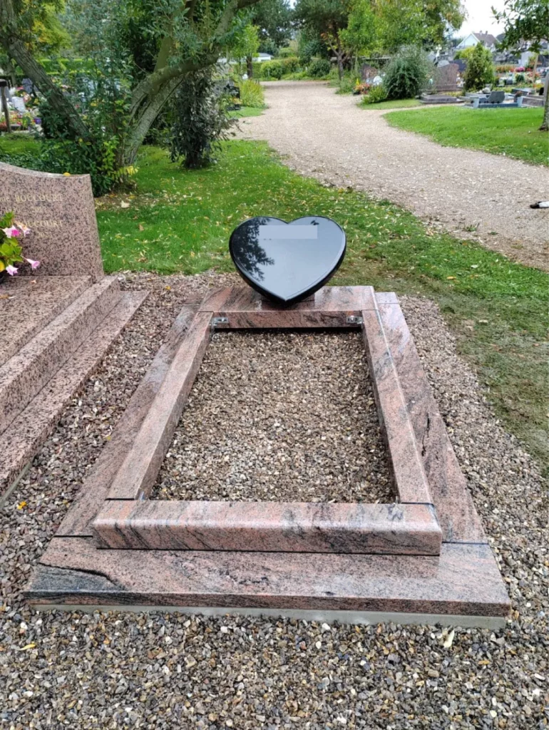 Monument funéraire en granit rose et noir en cours d'installation avec stèle en forme de cœur sur lit de graviers.